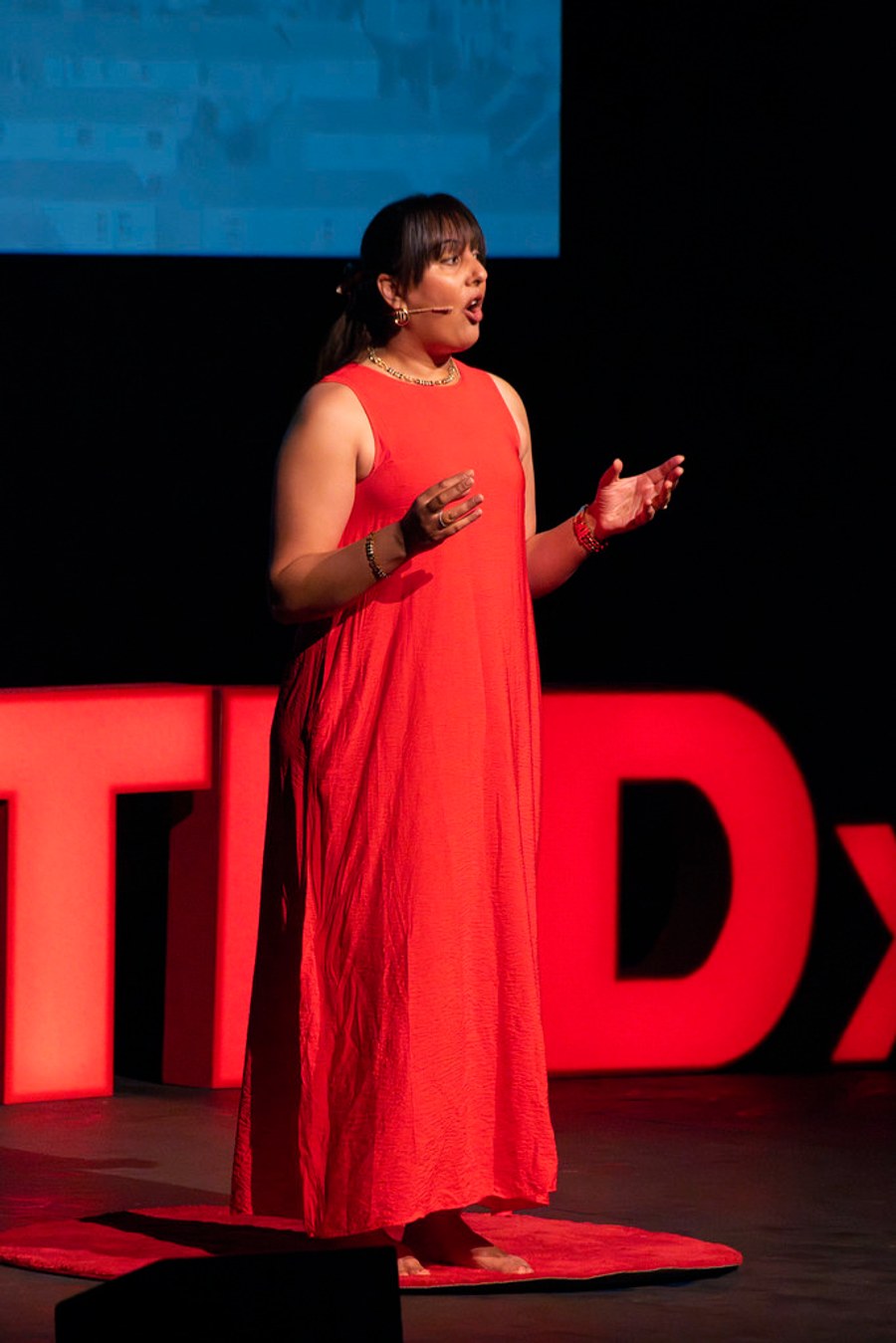 Nikita Thakrar dancing barefoot during her TEDxTruro talk, in a red dress, with the illuminated TEDx letters behind her