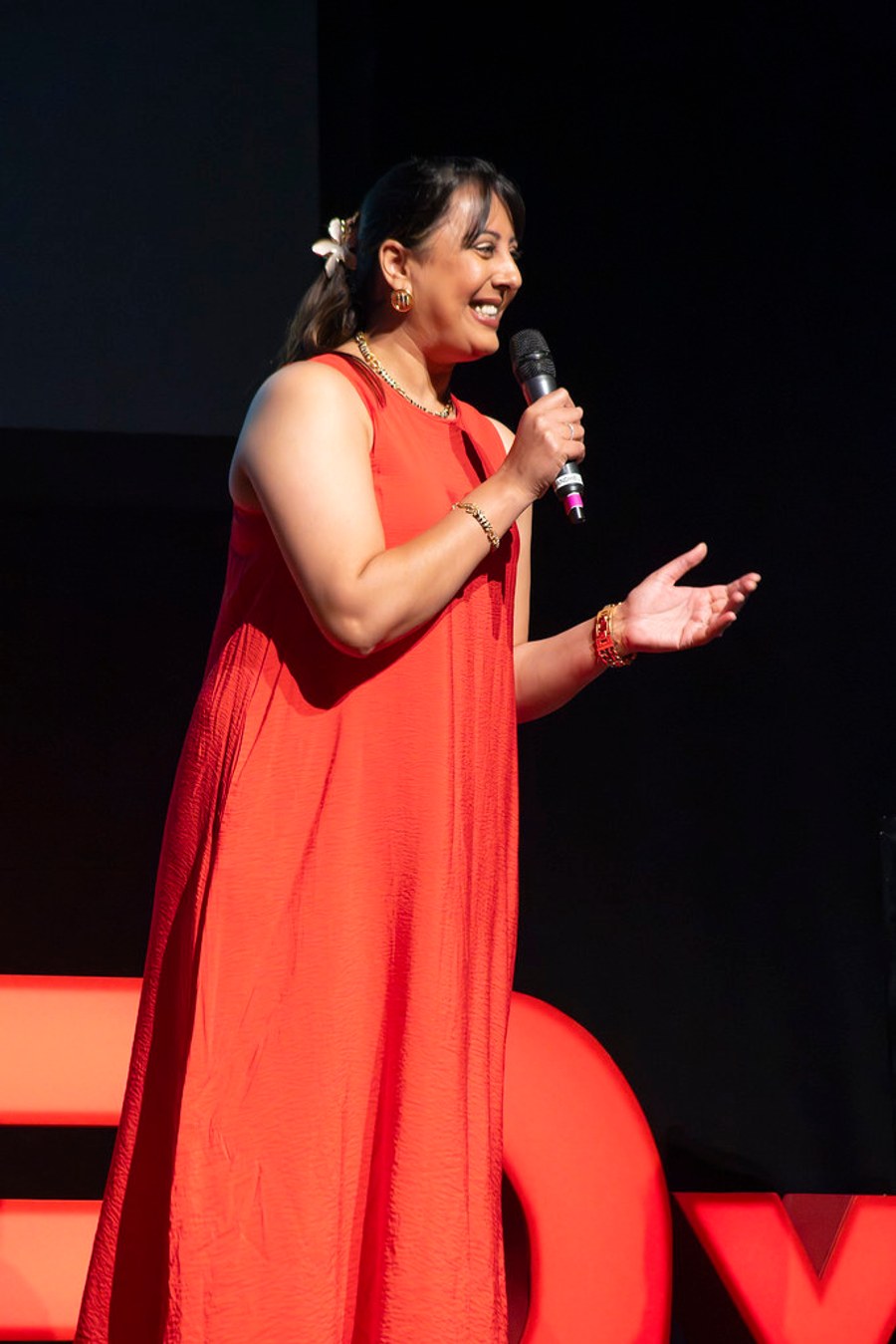 Nikita Thakrar speaking into a microphone on the TEDxTruro stage in a red dress